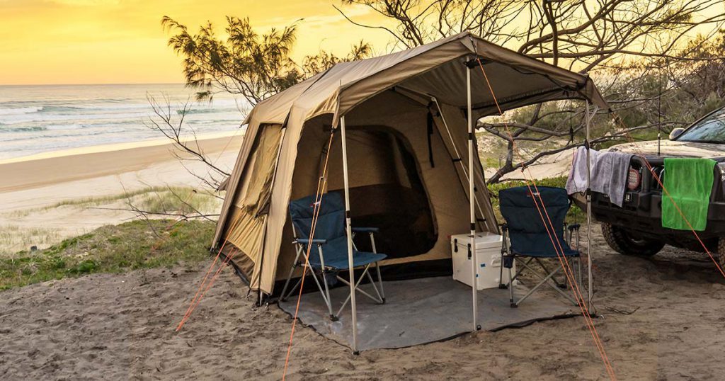 A tent and campsite scene by the ocean with a golden sunset behind.