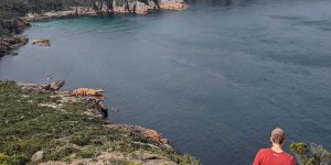Fraser is standing on the edge of a cliff, patchy with green shrubbery, overlooking the vast ocean below. He wears a red t-shirt, beige cap, and grey shorts. His back is to the camera, and the cliff stretches further around, visible in the background.