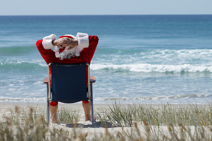 Santa sitting with his back to the camera in a beach chair, hands behind his head, watching the waves crash on the shore on a sunny day. 