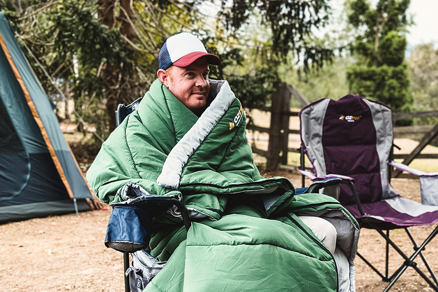 A man sitting in a camp chair, wrapped up in a sleeping bag, at a campsite. 
