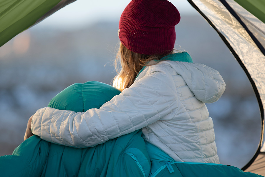 A woman wrapped up in a blue sleeping bag, wearing a red beanie, looking out beyond her tent. 