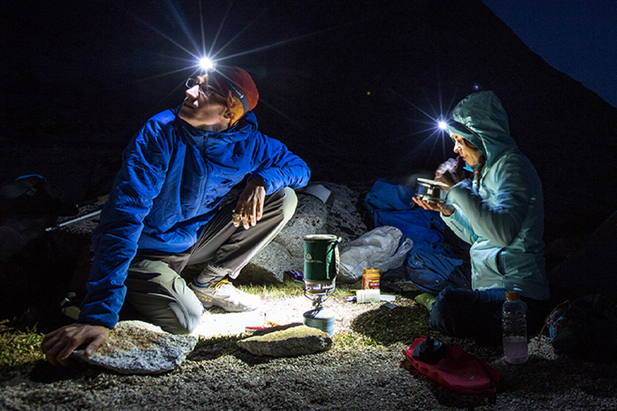 Our gear gurus shed light on choosing the perfect hands-free lighting solution for your outdoor adventures. Two campers cooking in the dark, both wearing headlamps.
