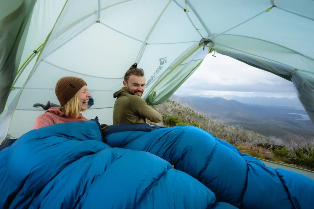 Couple in sleeping bags sitting inside a hiking tent with a valley view out the door.