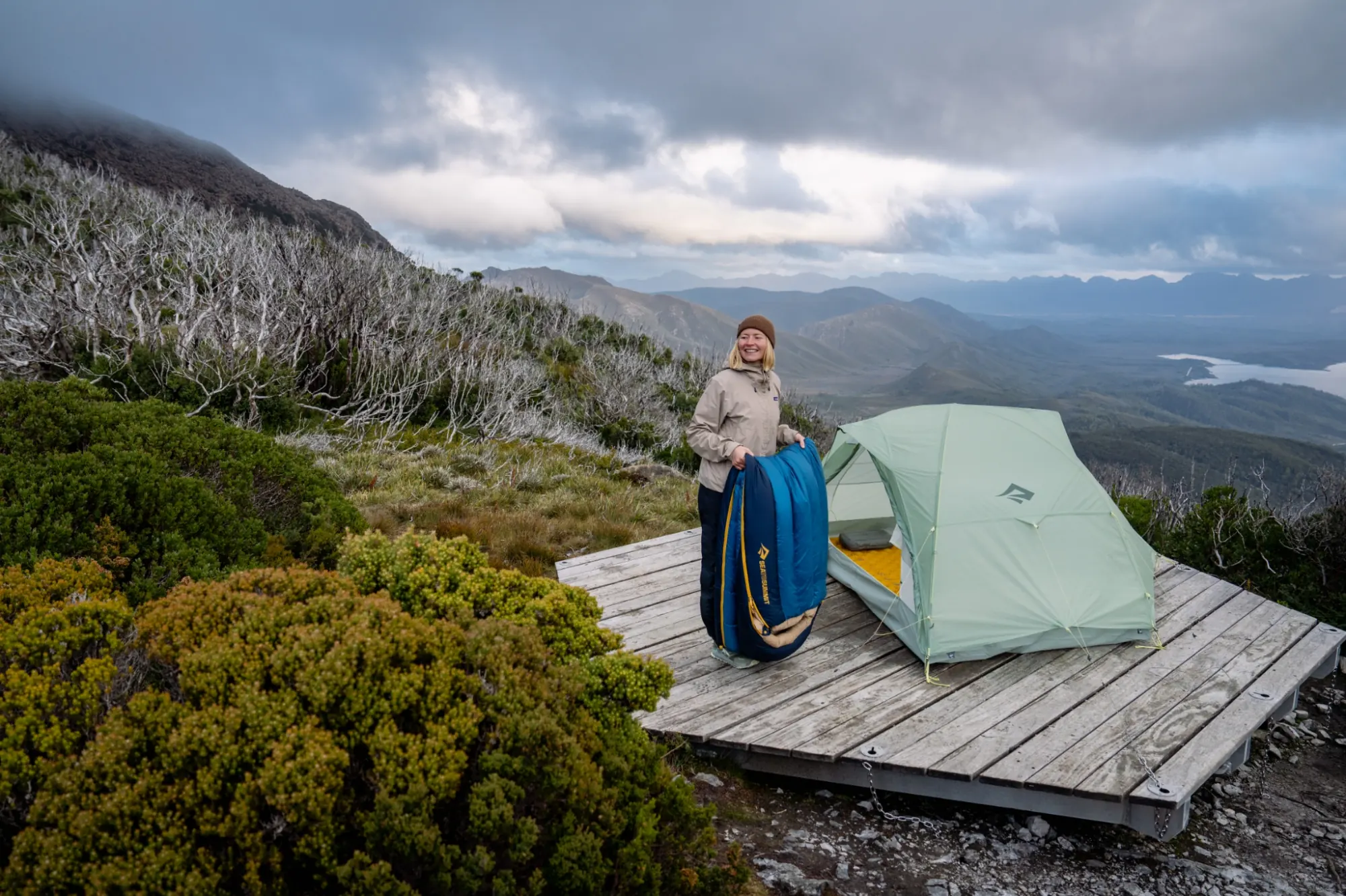 Our customers have made the decisions! Female standing next to a hiking a tent setup on a platform whilst holding a sleeping bag in remote wilderness scene