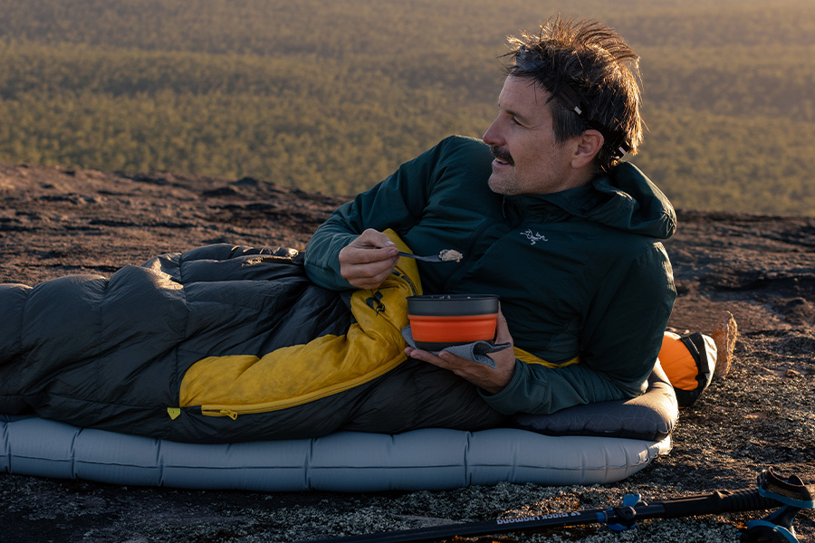 Ben and Lauren roll out the welcome mat for their all-time favourite guest, Ross from Sea to Summit A man snuggled up in a Sea to Summit sleeping bag atop a sleeping mat, eating breakfast from a bowl, on the edge of a cliff at sunrise.