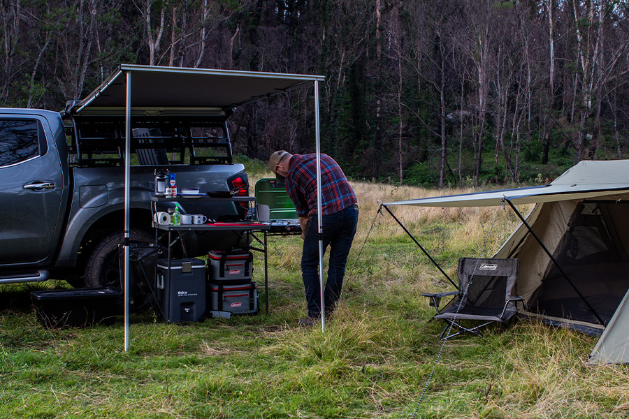 A man is setting up his camp kitchen from the side of his 4WD under an awning, with a tent and chair erect behind him. 