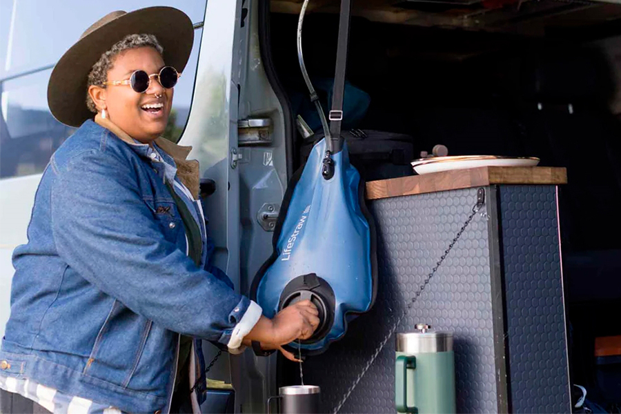 A go-getting, geared-up guide for anyone who's as ready to get out there as they are frozen at the front door! A camper wearing sunglasses, a hat, and a blue jacket smiling while filling their travel cup with water from a LifeStraw pouch.