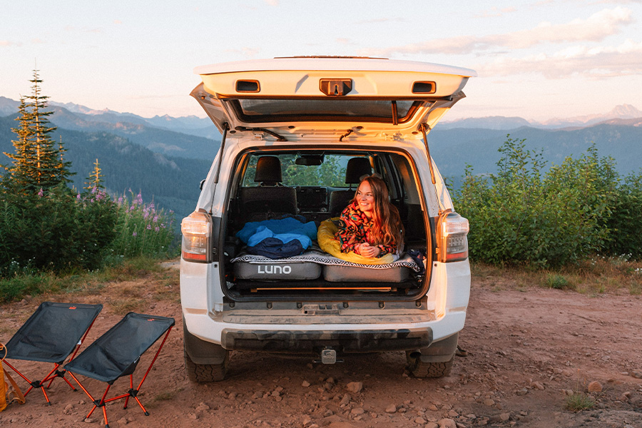 Tune in for essential, go-to guidance that reveals your ride's role in camping adventures A woman lies on her front in the back of her vehicle with the back lifted, on top of two Luno mattresses, smiling in the sunset on the mountaintop.