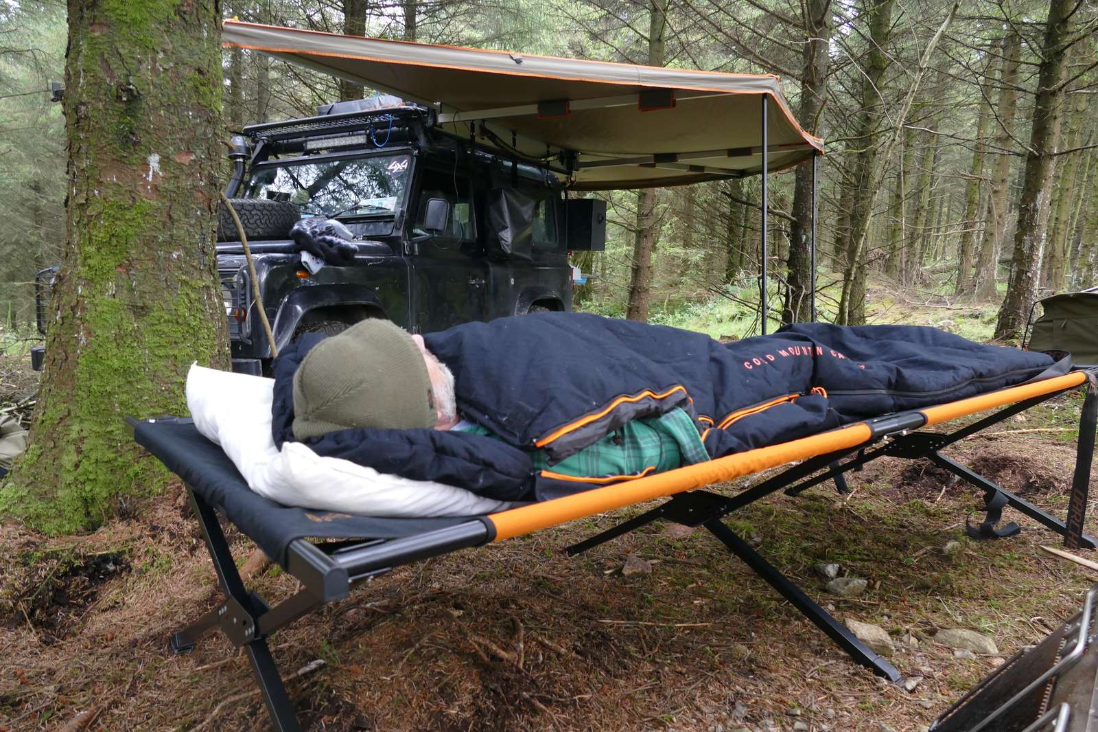 Camper sleeping on a stretcher in a sleeping bag next to a 4WD in the forest.